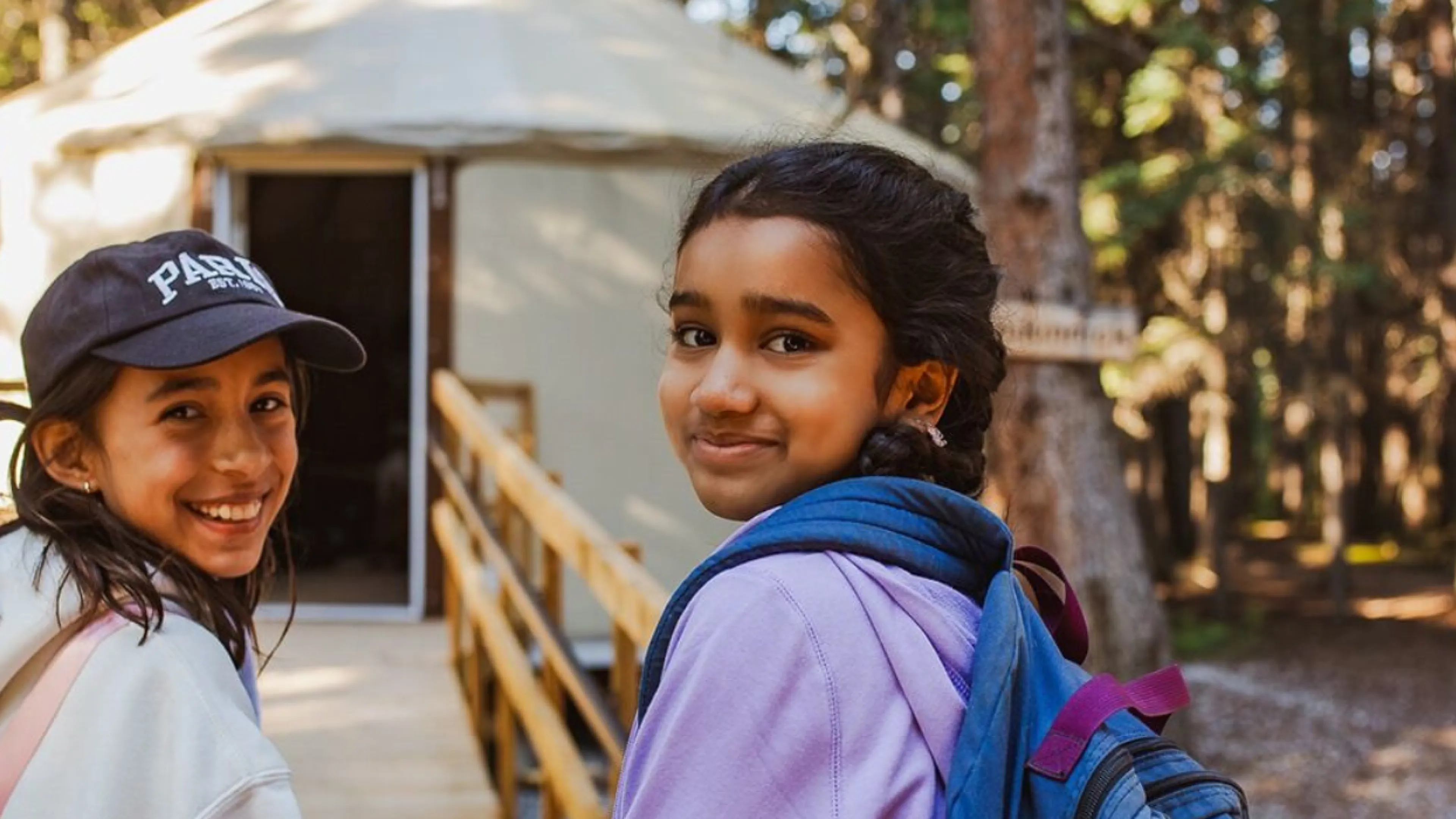 Two girls smiling in front of a yurt at camp in sunlight.